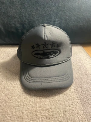 Grå truckerkeps med stjärnor - Snygg grå truckerkeps med svart broderad logga framtill och fem stjärnor ovanför. Kepsen har mesh på sidorna och bak för extra ventilation. Justerbar passform med snapback-spänne. Perfekt för en avslappnad streetstyle.