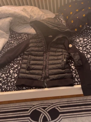 Svart pufferjacka från Moncler - Snygg svart pufferjacka från Moncler med quiltad front och huva. Ärmarna är i mjukt tyg och jackan har två stora fickor med dragkedja. Klassisk Moncler-logga på ena ärmen. Perfekt för kalla vinterdagar.