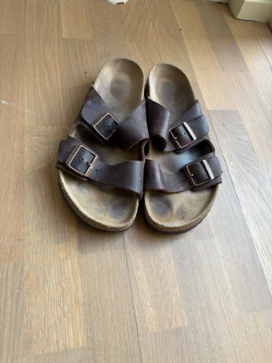 Bruna Birkenstock sandaler i läder - Klassiska bruna sandaler från Birkenstock med två breda remmar i läder och justerbara metallspännen. Fotbädden är anatomiskt utformad för extra komfort. Perfekta för en avslappnad och stilren look.