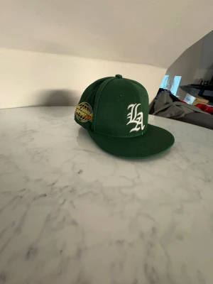 Grön LA keps med brodyr - Snygg grön keps med platt skärm och broderad LA-logga i vitt framtill. På sidan finns ett gult och blått broderat märke med texten 'Los Angeles Area Code 1988'. Klassisk streetwear-stil, perfekt för dig som gillar sportiga accessoarer.