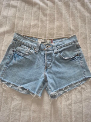 Levi's ljusblå jeansshorts low slouch - Ljusblå jeansshorts från Levi's med låg midja och fransiga benslut. Köpta på Vinted men som inte satt helt bra vilket gör att  jag säljer dessa vidare. Midjebredd är 38 cm och total längd 25 cm fram och 28 cm bak.