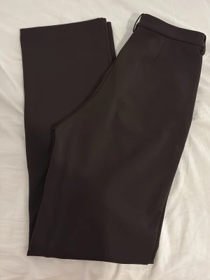 Bruna kostymbyxor från Vero Moda - Snygga bruna kostymbyxor från Vero Moda med raka ben och klassisk passform. Byxorna har hällor för skärp och sidfickor. Perfekta för en stilren look. Materialet är slätt och känns bekvämt mot huden.