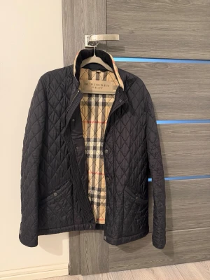 Svart quiltad jacka från Burberry Brit - Stilren svart quiltad jacka från Burberry Brit med klassiskt rutigt foder i beige, svart och rött. Jackan har knappar framtill, ståkrage och två fickor med lock. Perfekt för dig som vill ha en tidlös och snygg jacka med exklusiv känsla.