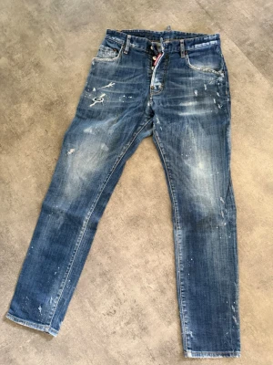 Blå jeans från Dsquared2 - Säljer ett par blå jeans från Dsquared2 med distressed detaljer, slitningar och färgstänk för en cool, trendig look. Jeansen har smal passform, klassisk femficksdesign och knappgylf. Perfekta för dig som gillar streetwear och unik stil.