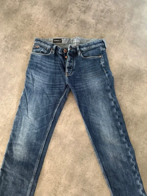 Blå jeans från Armani  - Säljer ett par klassiska blå jeans från Armani Jeans med snygg tvätt och raka ben. Jeansen har fyra fickor, kopparfärgade knappar och kontrastsömmar. Perfekta för dig som gillar en tidlös och avslappnad stil.