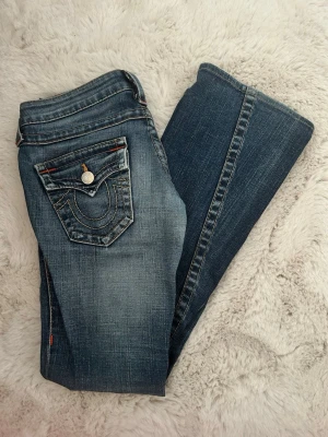 Blå bootcut jeans från True Religion - Snygga blå bootcut jeans från True Religion med klassisk kontrastsöm och ikonisk fickdesign bak. Jeansen har slitningar och detaljer i orange och silverfärgade knappar. Perfekta för dig som gillar en avslappnad men trendig look.