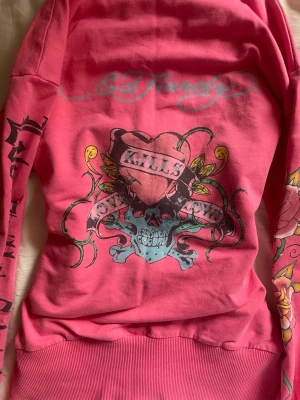 Skitttt cool rosa Ed Hardy hoodie!! - Säljer en rosa hoodie från Ed Hardy med dragkedja och huva. Hoodien har stora färgglada tryck med dödskalle, hjärta och texten 'Love Kills Slowly' på ryggen och ärmarna. Materialet är mjukt bomullstyg och modellen är klassisk streetwear. Super cool o perfekt för sommaren!!! Står storlek XL på den men passar som S/M för storlekarna är mkt mkt mindre😋❤️❤️