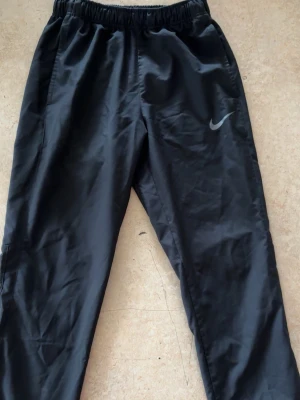 Svarta träningsbyxor från Nike - Svarta träningsbyxor från Nike med klassisk logga på vänster ben. Byxorna har elastisk midja och är tillverkade i ett lätt och glansigt syntetmaterial som passar perfekt för sport och aktivitet. Snygg och enkel design som är lätt att matcha med andra plagg.