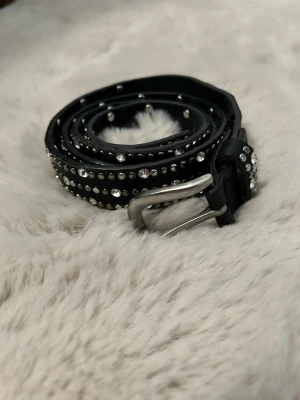 Svart bälte med nitar och strass - Snyggt svart bälte i skinn med silverfärgat spänne. Bältet är dekorerat med rader av små silvernitar och glittrande strassdetaljer som ger en cool och trendig look. Perfekt accessoar för att lyfta din outfit.