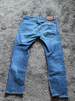Blå raka jeans från Replay - Säljer ett par blå jeans från Replay med klassisk femficksdesign och raka ben (Rocco). Jeansen har snygga detaljer på bakfickorna och en läderpatch med logga bak i midjan. Materialet är slitstark denim som känns bekväm. Nypris 1499kr.