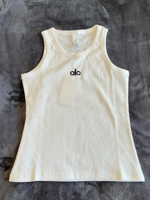 Vit ribbad linnetopp från Alo - Supersnygg vit ribbad linnetopp från Alo med broderad logga framtill. Toppen är ärmlös och har en klassisk rund halsringning. Perfekt för en sportig och trendig look.