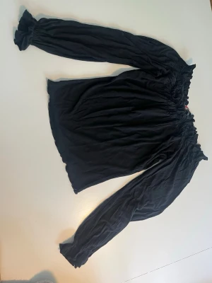 Svart offshoulder-blus med resår - Trendig svart offshoulder-blus med långa ärmar och resår vid halsringning, ärmslut och nederkant. Blusen har en avslappnad passform och är tillverkad i ett mjukt, lätt tyg som ger en snygg siluett.