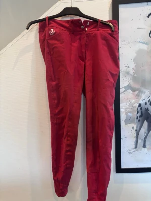 Röda ridbyxor från Jacson - Snygga röda ridbyxor från Jacson med logotyp på fickan. Byxorna är tillverkade i en mix av bomull, nylon och lycra vilket gör dem både bekväma och stretchiga. De har knäppning framtill och smal passform som sitter tight längs benen.