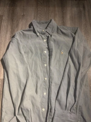 Skjorta från Polo Ralph Lauren - Klassisk grå skjorta från Polo Ralph Lauren med knappar framtill och broderad logga på bröstet. Skjortan har lång ärm och en tidlös design som passar till många olika stilar. Storlek M