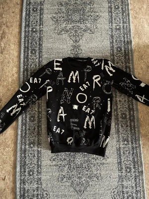 Svart sweatshirt EA7 Emporio Armani - Cool svart sweatshirt från EA7 Emporio Armani med vit grafisk text och logga över hela tröjan. Rund halsringning och långa ärmar. Perfekt för en trendig streetwear-look.