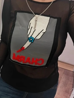 Svart mesh topp med CHARM-tryck - Snygg svart mesh topp från Vince Camuto med lång ärm och stort tryck framtill av en hand med blå ring och röd texten 'CHARM'. Toppen är genomskinlig och har en cool, edgy vibe som passar perfekt till jeans eller kjol.