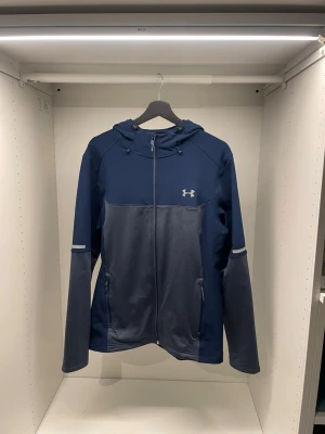Blå träningsjacka från Under Armour - Snygg blå träningsjacka från Under Armour med huva och dragkedja. Jackan har två fickor med dragkedja, reflekterande detaljer på ärmarna och logga på bröstet. Perfekt för utomhusträning och sportiga aktiviteter. Kvitto finns.