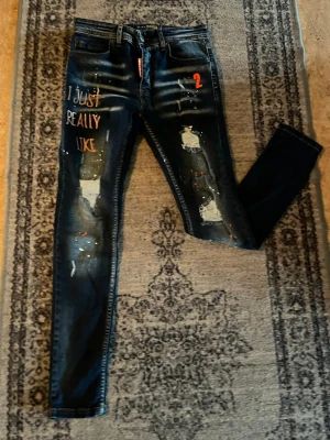 Mörkblå skinny jeans med tryck - Unika mörkblå skinny jeans med slitningar, färgstänk och texten 'I JUST REALLY LIKE' på vänster ben. Jeans med patchade hål och orange detaljer, perfekt för dig som vill sticka ut med din stil.