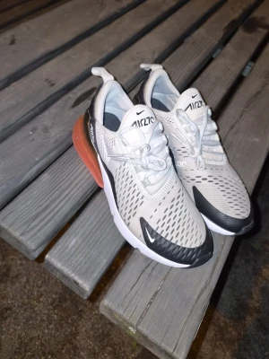 Nike Air Max 270 vita sneakers - Nike Air Max 270 sneakers i vitt med svarta detaljer och en tydlig orange Air Max-sula. Ovandelen är i mesh för bra andningsförmåga och har klassiska Nike-loggor på plösen och sidan. Snygg och sportig design med snörning och låg profil.
