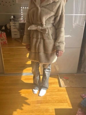 Beige fuskpäls kappa - Mysig beige kappa i fuskpäls med huva och knytskärp i midjan. Kappan har en fluffig och lyxig känsla, perfekt för kyliga dagar. Lång modell som går ner över låren och har breda ärmar. Snygg och trendig look. De står för den som köper att betala frakten