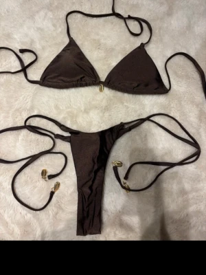 Brun bikini med gulddetaljer - Snygg brun bikini med trekantsöverdel och smala knytband. Underdelen har hög skärning och båda delarna har fina guldfärgade detaljer på banden. Perfekt för stranden eller poolen.