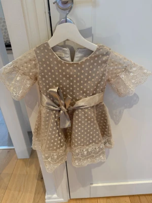 Söt beige prickig klänning med rosett - Beige kortklänning med prickigt mönster, spetsdetaljer och en bred satinrosett i midjan. Klänningen har korta ärmar med spets och rund halsringning. Perfekt för speciella tillfällen och har en elegant känsla.