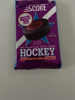 Score NHL Hockeykort 1993-94 oöppnat - Oöppnat paket med 15 hockeykort från Score, säsongen 1993-1994 NHL. Lila och blå förpackning med text och grafik. Innehåller 14 spelarkort och 1 bonus Pinnacle-kort. Perfekt för samlare av sportkort och hockeymemorabilia.