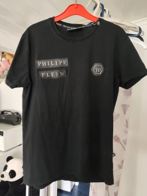 Svart Philipp Plein t-shirt - Snygg svart t-shirt från Philipp Plein med logotypdetaljer på bröstet. Klassisk rund halsringning och korta ärmar. Tillverkad i mjuk bomull för en bekväm känsla. Perfekt för dig som gillar streetwear och vill sticka ut med ett exklusivt märke.