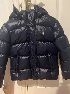 Mörkblå pufferjacka från Polo Ralph Lauren - Snygg mörkblå pufferjacka från Polo Ralph Lauren med huva och klassisk logga på bröstet. Jackan har stora fickor framtill och en glansig finish. Perfekt för kalla vinterdagar och riktigt varm tack vare den tjocka vadderingen.