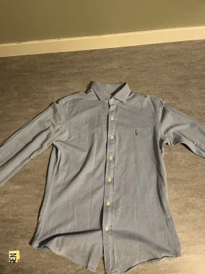Blå skjorta från Ralph Lauren - Klassisk ljusblå skjorta från Ralph Lauren med broderad logga på bröstet. Skjortan har lång ärm, knappar hela vägen och en stilren krage. Perfekt till jeans eller chinos för en clean look.