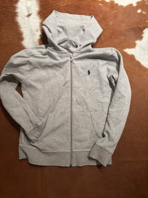 Grå hoodie från Polo Ralph Lauren - Snygg ljusgrå hoodie med dragkedja från Polo Ralph Lauren. Klassisk design med huva, två fickor framtill och den ikoniska lilla broderade loggan på bröstet. Perfekt för en chill och stilren look.