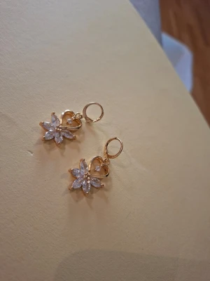 Guldiga örhängen med blomform - Snygga örhängen i guldfärg med hängande blomformade hängen dekorerade med gnistrande stenar. Blommorna har sex blad och örhängena har en liten ring som fäste. Perfekta för att ge din look lite extra glans.