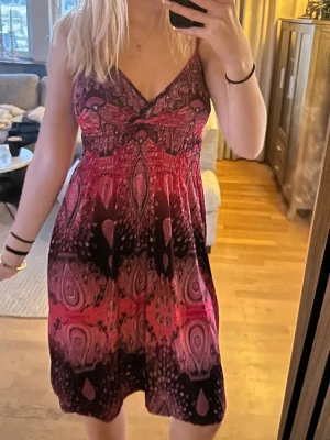 Rosa och svart mönstrad sommarklänning - Säljer en kort klänning med smala axelband och v-ringning. Klänningen har ett färgstarkt mönster i rosa och svart med inslag av lila och har en avslappnad passform. Perfekt för varma dagar och enkel att styla med sandaler eller sneakers.