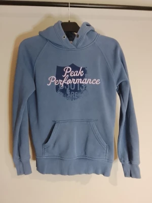 Blå hoodie från Peak Performance - Snygg blå hoodie från Peak Performance med tryckt logga i ljus rosa på bröstet (syns inte bra på bilden men jättefin ljusrosa är hela texten på bröstet). Klassisk känguruficka framtill och ribbade muddar. Mjuk insida och huva med dragsko. Perfekt för en avslappnad och sportig stil. Tröjan är i mycket bra skick, den ska inte vara "ljus lila" till färgen så den är inte "sliten" i färgen utan ska vara så, riktigt fin men tyvärr passar den inte mig. Äkta såklart. Inköps pris 1899kr