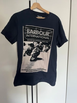 Svart Barbour International t-shirt - Cool svart t-shirt från Barbour International med stort tryck av motorcyklar och logga på framsidan. Klassisk rund hals och korta ärmar. Perfekt för dig som gillar streetwear och snygga prints.