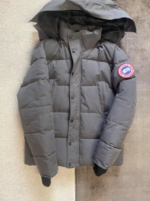 Grå dunjacka från Canada Goose - Säljer en grå dunjacka från Canada Goose med klassisk patch på ärmen. Jackan har huva, hög krage och knäppning med både dragkedja och knappar. Fylld med dun för extra värme och har en stilren, boxig passform.