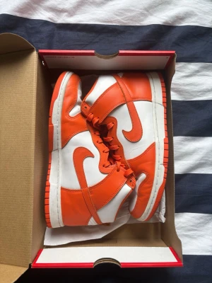 Nike Dunk High Orange/White sneakers - Nike Dunk High sneakers i orange och vitt läder med klassisk Swoosh-logga på sidorna. Skorna har högt skaft, matchande orange snörning och vit mellansula. Perfekt för dig som gillar streetwear och vill sticka ut med färg.