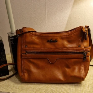Brun axelväska från Mandarina Duck - Snygg brun axelväska i slätt konstläder från Mandarina Duck. Väskan har justerbar axelrem, flera dragkedjefack framtill och en stilren design med diskret logga. Perfekt för dig som vill ha en praktisk och trendig väska till vardagslooken.
