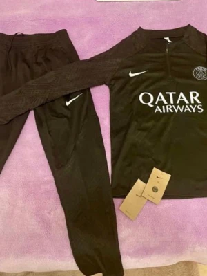 Svart PSG träningsset från Nike - Säljer ett svart träningsset från Nike och PSG med långärmad tröja och byxor. Tröjan har halv dragkedja, PSG-logga och Qatar Airways-tryck på bröstet. Byxorna har Nike-logga och smal passform. Perfekt för fotbollsträning eller chill.