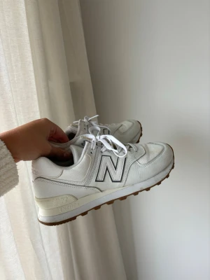 Vita New Balance sneakers 574 - Säljer ett par klassiska vita New Balance 574 sneakers med mesh och läderdetaljer. Skorna har en stor N-logga på sidan, vita snören och en brun yttersula med ENCAP-dämpning. Perfekt för dig som gillar stilrena och sportiga sneakers.