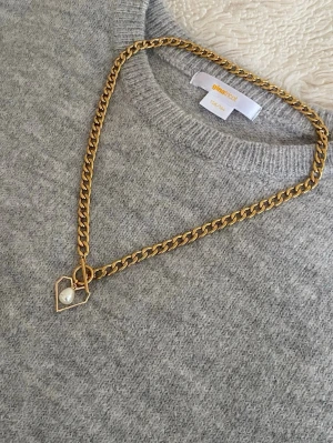 Guldfärgat halsband med hjärthänge - Stilrent halsband i guldfärgad rostfritt med en kedja i chunky modell. Hänget är format som ett hjärta med en vit pärldetalj i mitten. Perfekt accessoar för att lyfta din outfit med en trendig touch.