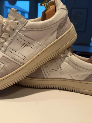 Hackett London Sneakers – stilrena premium-sneakers i vitt läder - Säljer ett par exklusiva sneakers från Hackett London, modell Dexter Winter / White Heritage-serien, i riktigt snygg och tidlös design.  ✔ Ovandel i vitt premiumläder ✔ Eleganta ljusgrå mockadetaljer runt tå och häl ✔ Karakteristiskt perforerat “H” på sidan ✔ Off-white/krämfärgad gummisula med modern räfflad struktur ✔ Stilren och lyxig sneaker som passar både vardag och smart casual  Hackett London är känt för sin brittiska premiumdesign där klassisk stil möter modern komfort. 