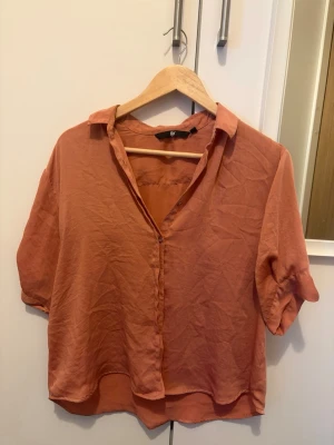 Rostfärgad blus från H&M - En snygg rostfärgad blus från H&M med avslappnad passform och krage. Blusen har knappar framtill och korta ärmar med lätt uppvik. Tillverkad i ett tunt, mjukt tyg som ger en luftig känsla.
