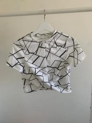 Vit topp med svart grafiskt mönster - Vit croppad topp med korta ärmar och svart grafiskt mönster. T-shirten har en rund halsringning och är tillverkad i ett mjukt material som känns bekvämt mot huden. Perfekt för en trendig och avslappnad stil.