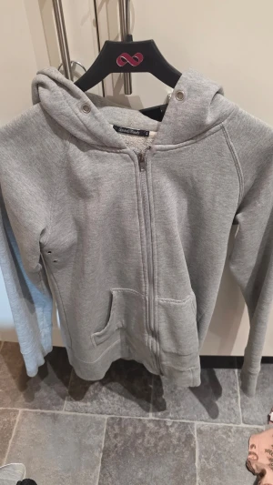 Grå zip-hoodie från Saints & Mortals - Grå hoodie med dragkedja från Saints & Mortals. Mjuk insida, huva med snörhål och två fickor framtill. Klassisk rak passform och långa ärmar. Perfekt för en avslappnad stil.