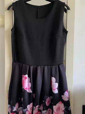 Svart klänning med rosa blommor - Snygg svart kortklänning med ärmlös design och rund halsringning. Kjoldelen har ett vackert blommigt mönster i rosa och vitt mot svart botten. Klänningen har en markerad midja och lätt utställd kjol. 