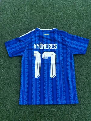 Blå Sverige fotbollströja Adidas - Blå fotbollströja från Adidas med Sverige-emblem och nummer 17 Gyökeres. Tröjan har ett unikt mönster i olika blå nyanser, vita detaljer och klassiska Adidas-ränder på axlarna. Perfekt för fotbollsfans som vill sticka ut på läktaren eller planen.