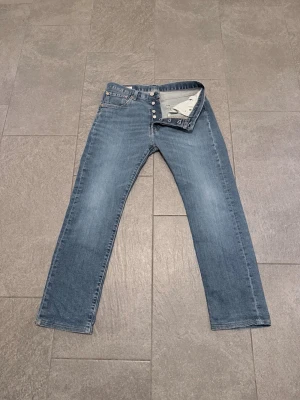 Blå raka Levi jeans - Den är upp syde men man ser måtten. Säljer ett par klassiska blå jeans med rak passform och knappgylf. Jeansen har fem fickor och detaljer i metall. Perfekta för en avslappnad och tidlös stil. Materialet är denim i bomull.