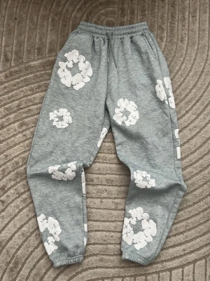 Grå Denim Tears mjukisbyxor  - Grå mjukisbyxor med vita blommönster över hela byxorna. Byxorna har resår i bensluten och en avslappnad passform. Perfekta för chill dagar hemma eller när du vill ha en skön och trendig look. Materialet känns mjukt och bekvämt mot huden.