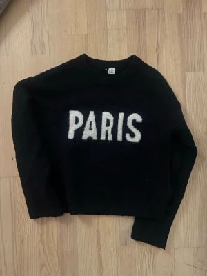 Svart stickad tröja med PARIS-tryck - Marinblå/svart Cool svart stickad tröja från Divided med stort vitt PARIS-tryck på bröstet. Tröjan har rund halsringning och långa ärmar. Perfekt för dig som gillar streetstyle och vill ha en statement-piece i garderoben.